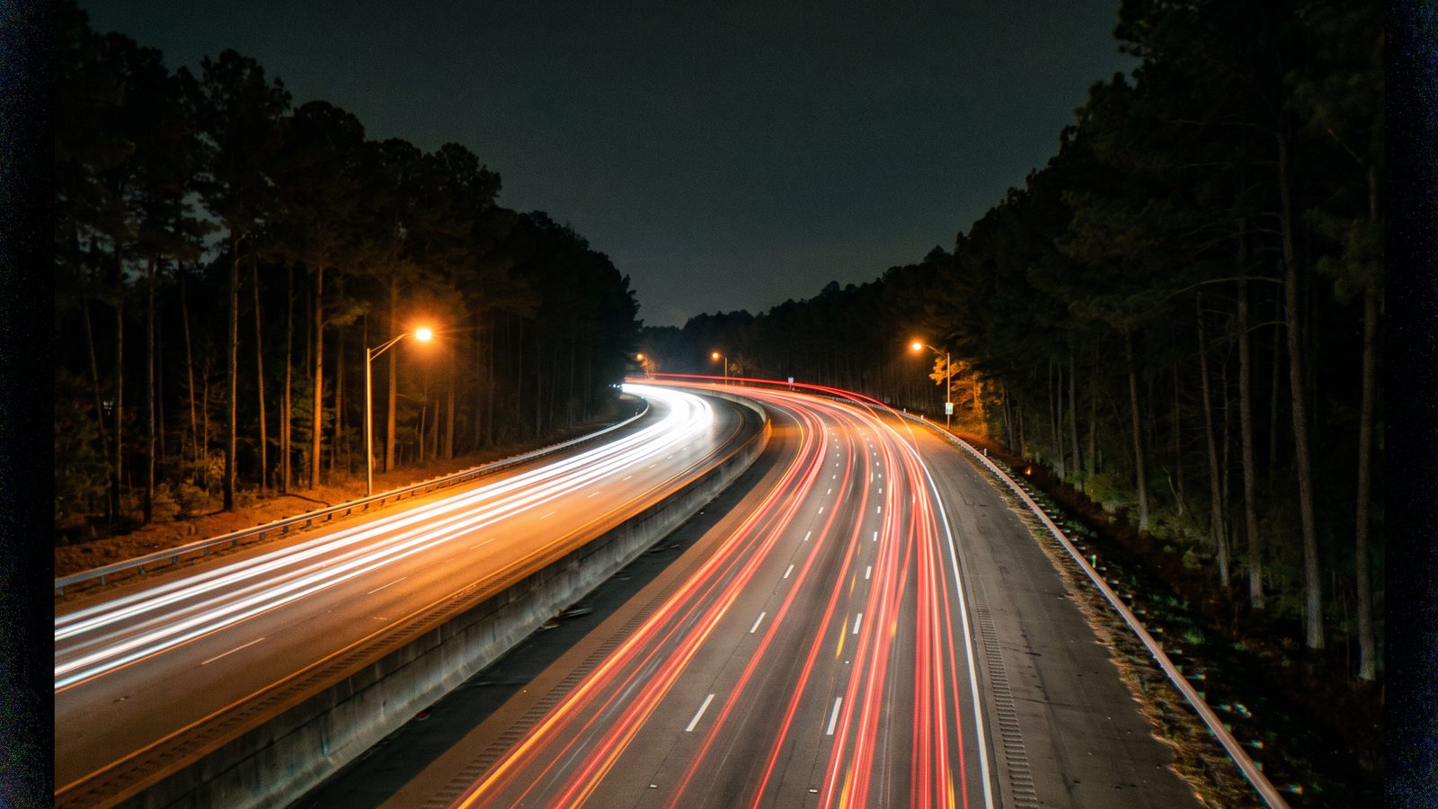 An interstate through pine woods at night with headlight and taillight trails (illustrative photo).